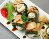Deliziosi involtini di zucchine grigliate ripieni di formaggio cremoso, guarniti con pomodorini, rucola e salsa scura.
