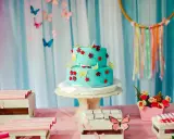 Torta a due piani decorata con farfalle e fiori in pasta di zucchero, su un tavolo rosa con palloncini rossi e caramelle.
