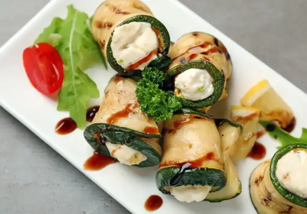 Deliziosi involtini di zucchine grigliate ripieni di formaggio cremoso, guarniti con pomodorini, rucola e salsa scura.