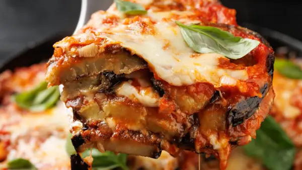 Una fetta di parmigiana di melanzane non fritte, con strati di melanzane, sugo e mozzarella filante, guarnita con basilico fresco.