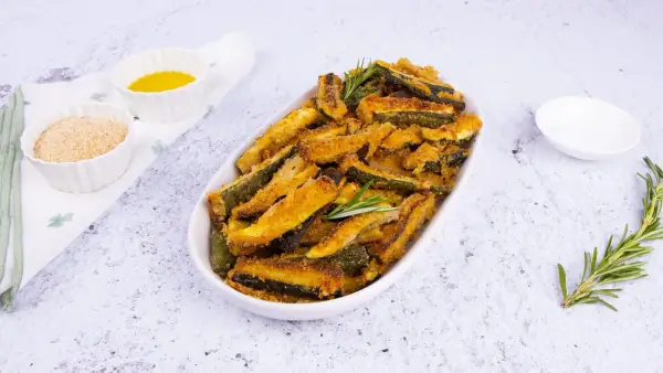 Zucchine gratinate al forno, dorate e croccanti, servite in un piatto ovale con rosmarino fresco. Accanto, ciotoline con pangrattato e olio.
