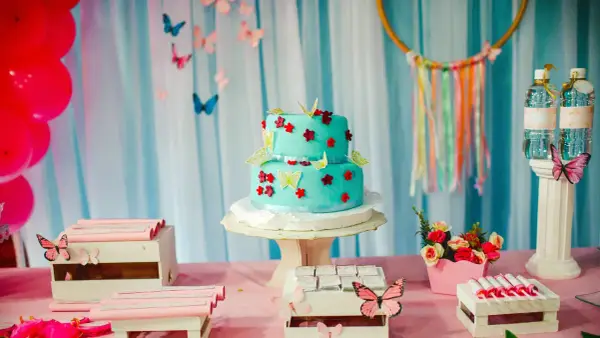 Torta a due piani decorata con farfalle e fiori in pasta di zucchero, su un tavolo rosa con palloncini rossi e caramelle.