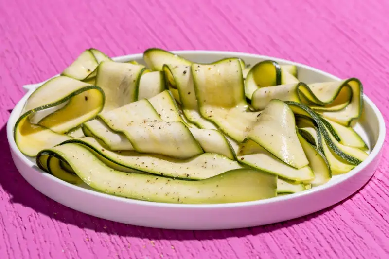 Fettine sottili di zucchine marinate crude, condite con pepe nero, su un piatto bianco, su un tavolo rosa acceso.