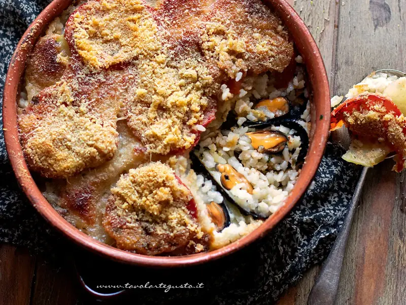 Piatto invitante di riso patate e cozze, gratinato con pomodoro e pangrattato, servito in una terrina di terracotta.